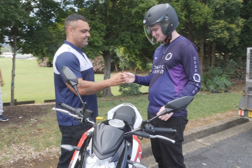 A man hands some keys to a younger man in a motorcycle helmet. 
