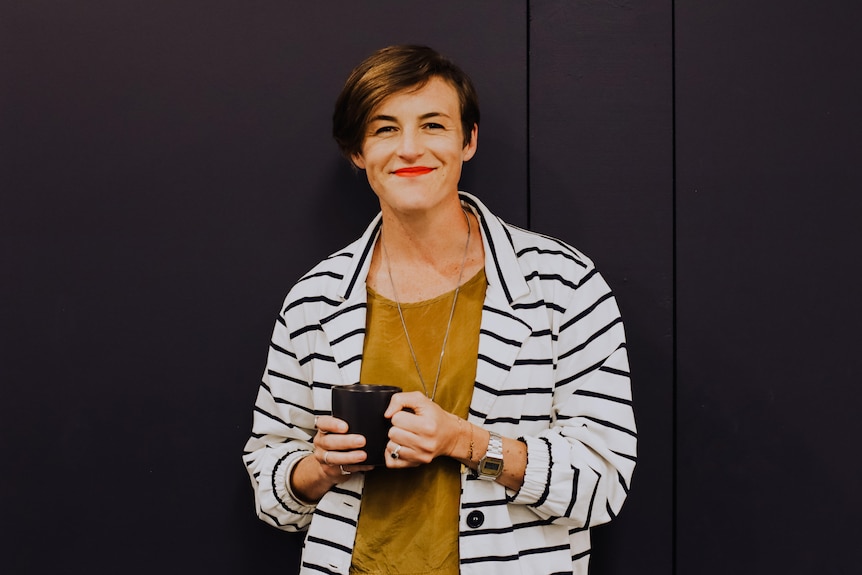 woman wearing a mustard shirt and striped jacket holds a mug and stands against a black door. A 