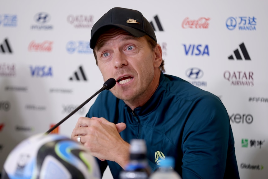 A cap-wearing Matildas coach speaks into a microphone while sitting at a press conference.