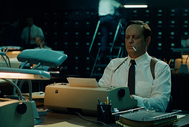 A man with grumpy expression in white shirt, suspenders and black tie sits at desk typewriter and cigarette in mouth in office.