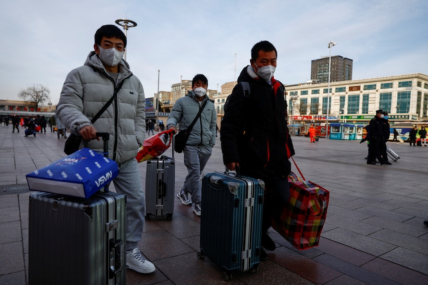 Three people in masks wheel their luggage outside.