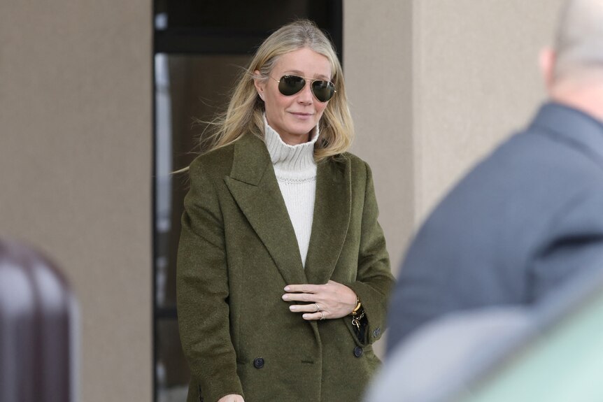 Gwyneth Paltrow in aviators and a green coat walks out of a building