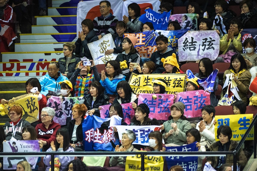 Middle-age women in a box at an ice rink clap and hold signs in expressing support for Yuzuru Hanyu
