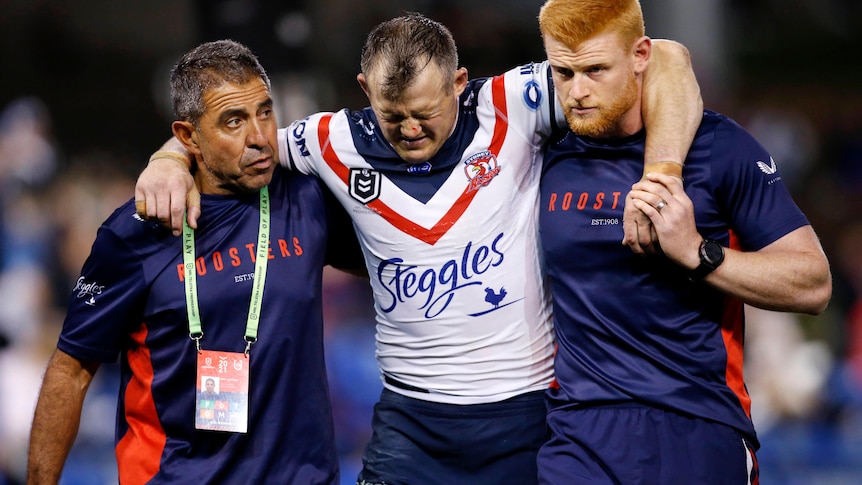 An NRL player is supported by two medics as he looks down and grimaces in pain. 