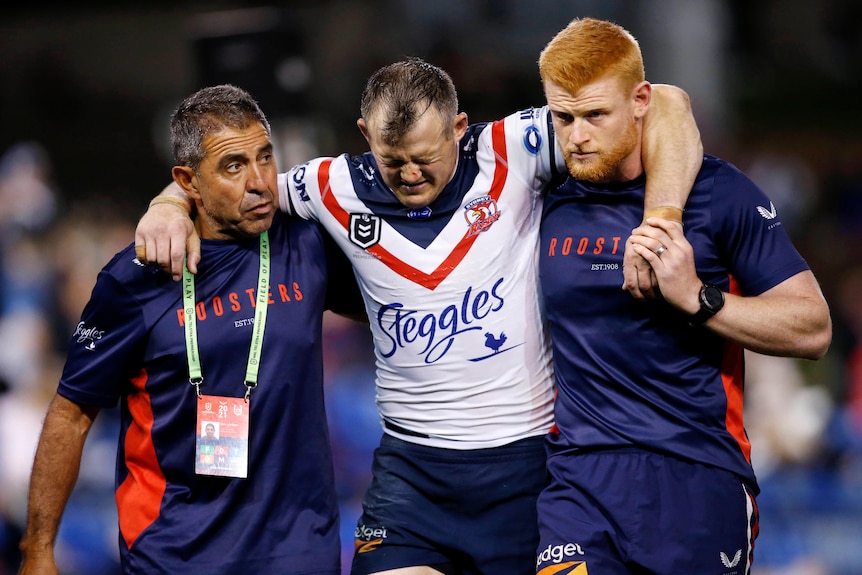 An NRL player is supported by two medics as he looks down and grimaces in pain. 