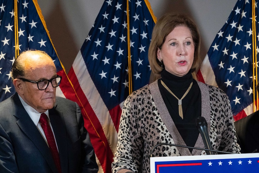 A woman is speaking at a podium and a man is standing next to her