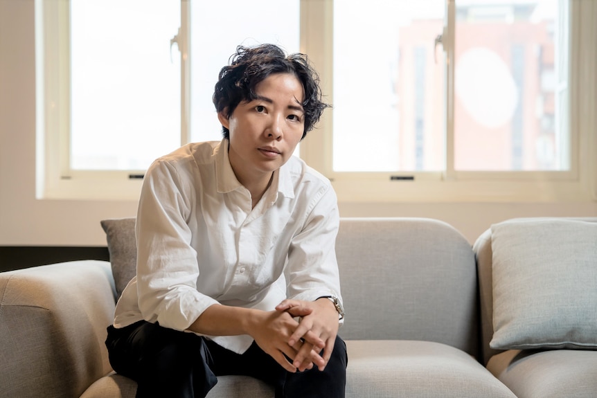 A Taiwanese woman with short hair in white buttoned shirt sitting on couch looking at camera.