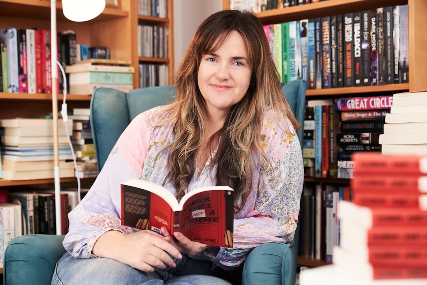 A woman with long highlighted brown hair sitting in a chair with bookshelves behind her, holding a book with a red cover open.