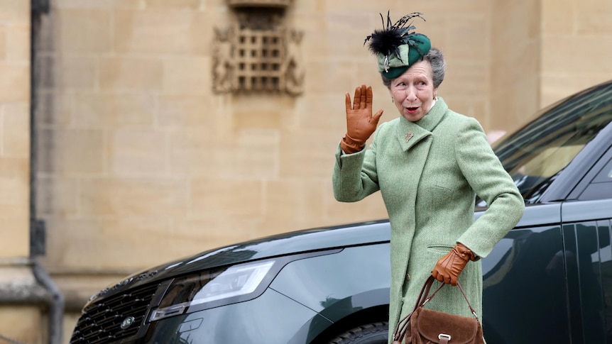 Princess Anne waves as she walks past a car. She wears a green coast and hat, brown gloves and holds a brown bag.