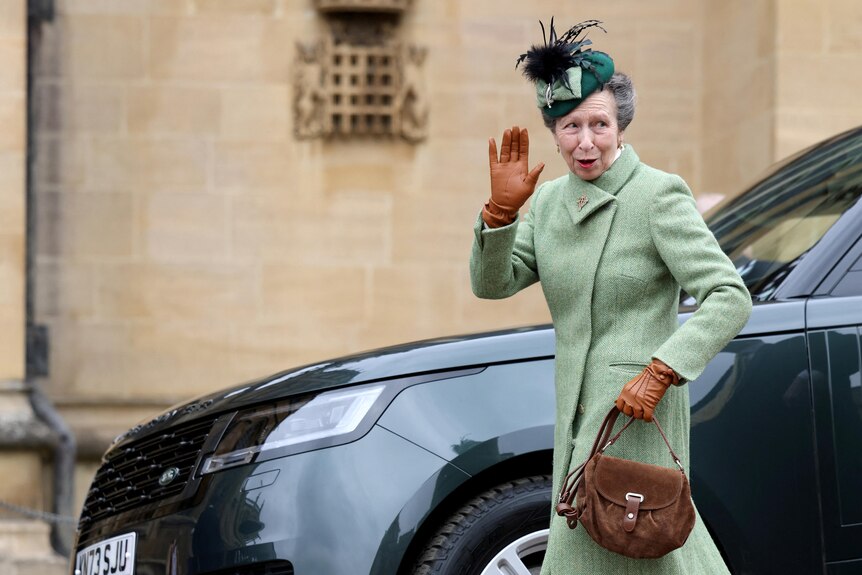 La princesa Ana saluda mientras pasa junto a un coche.  Lleva una costa verde y un sombrero, guantes marrones y sostiene una bolsa marrón.