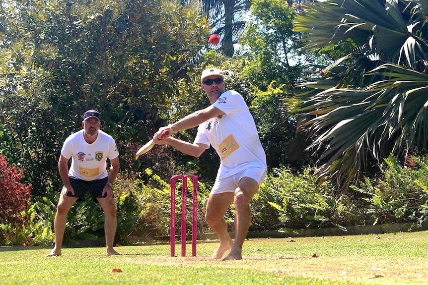 A man wearing cricket whites and cap swinging a cricket bat at a red ball.