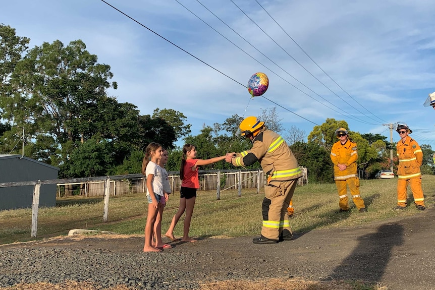 A firefighters hands a young girl a balloon while she stands with two friends.