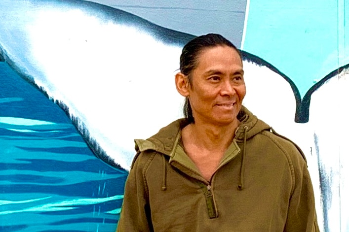 A man in long-sleeves and combat pants stands smiling, leaning back against a wall mural featuring a whale tail.  