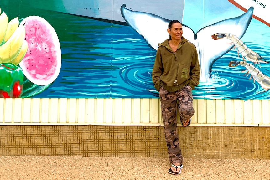 A man in long-sleeves and combat pants stands smiling, leaning back against a wall mural featuring a whale tail.  