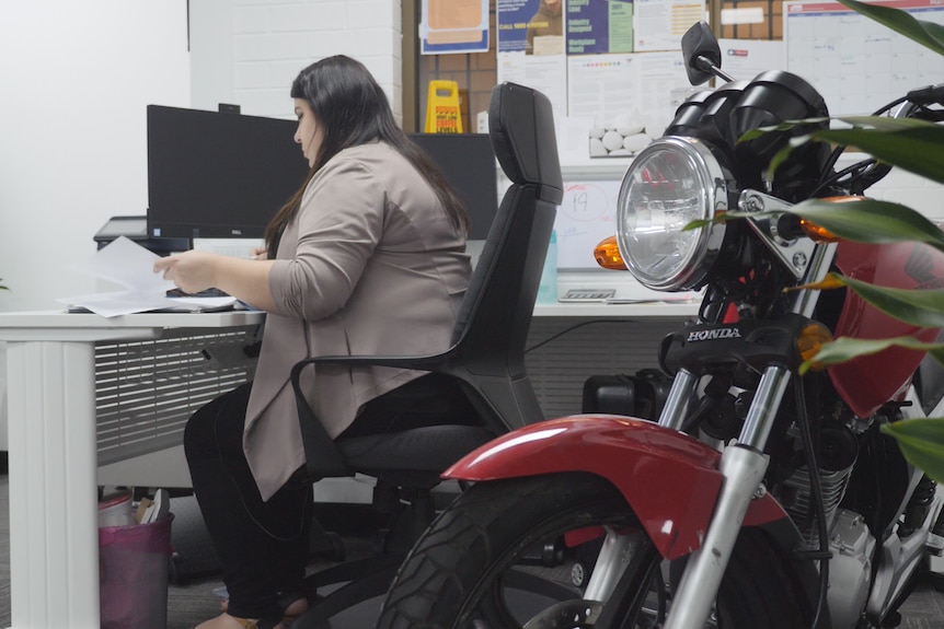 A woman sits at a computer, with a motorbike in the office.