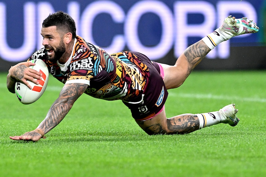 A Brisbane Broncos NRL player dives with the ball in his right hand to score a try.