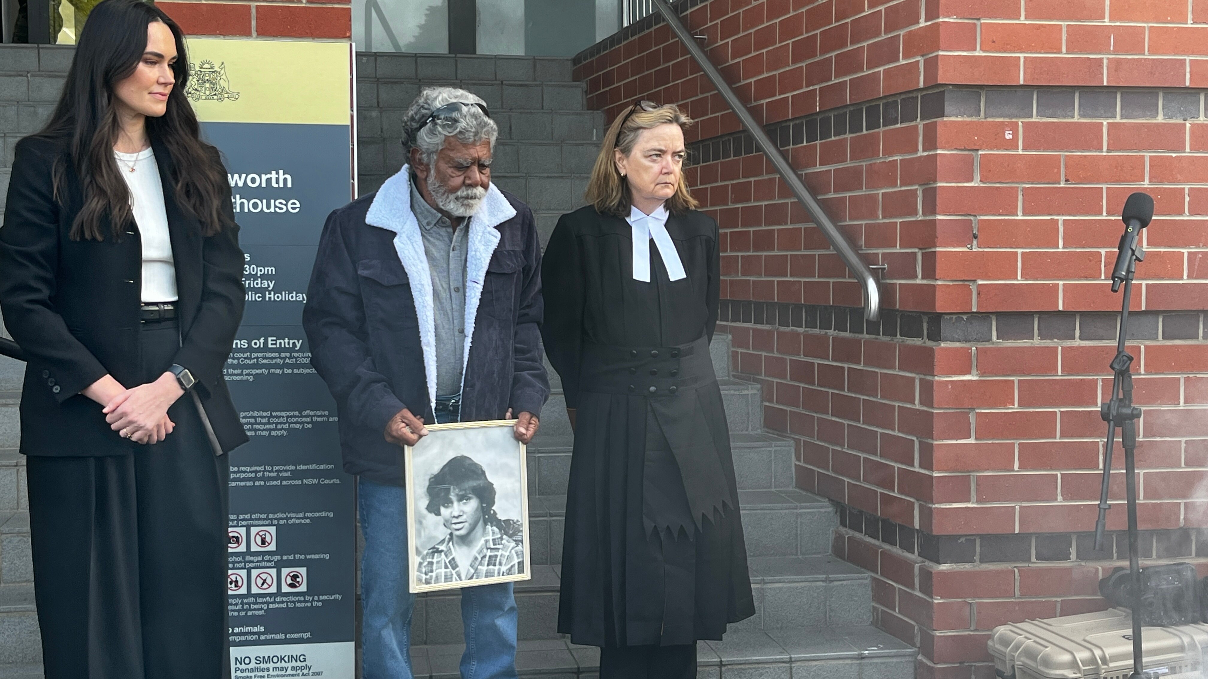 An Indigenous man with grey hair and beard holds a picture as he stands beside a judge in robes.