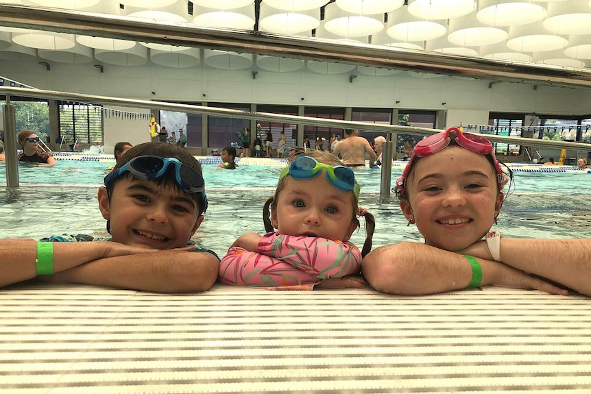 Three kids smiling on the side of an indoor public pool