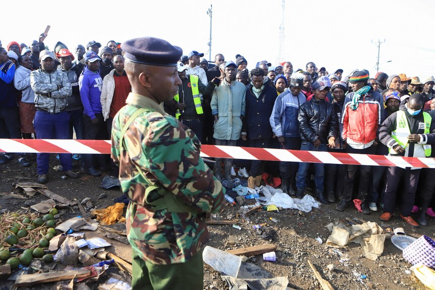 A Kenyan man in army gear stands at site of crash with people standing behind tape. 