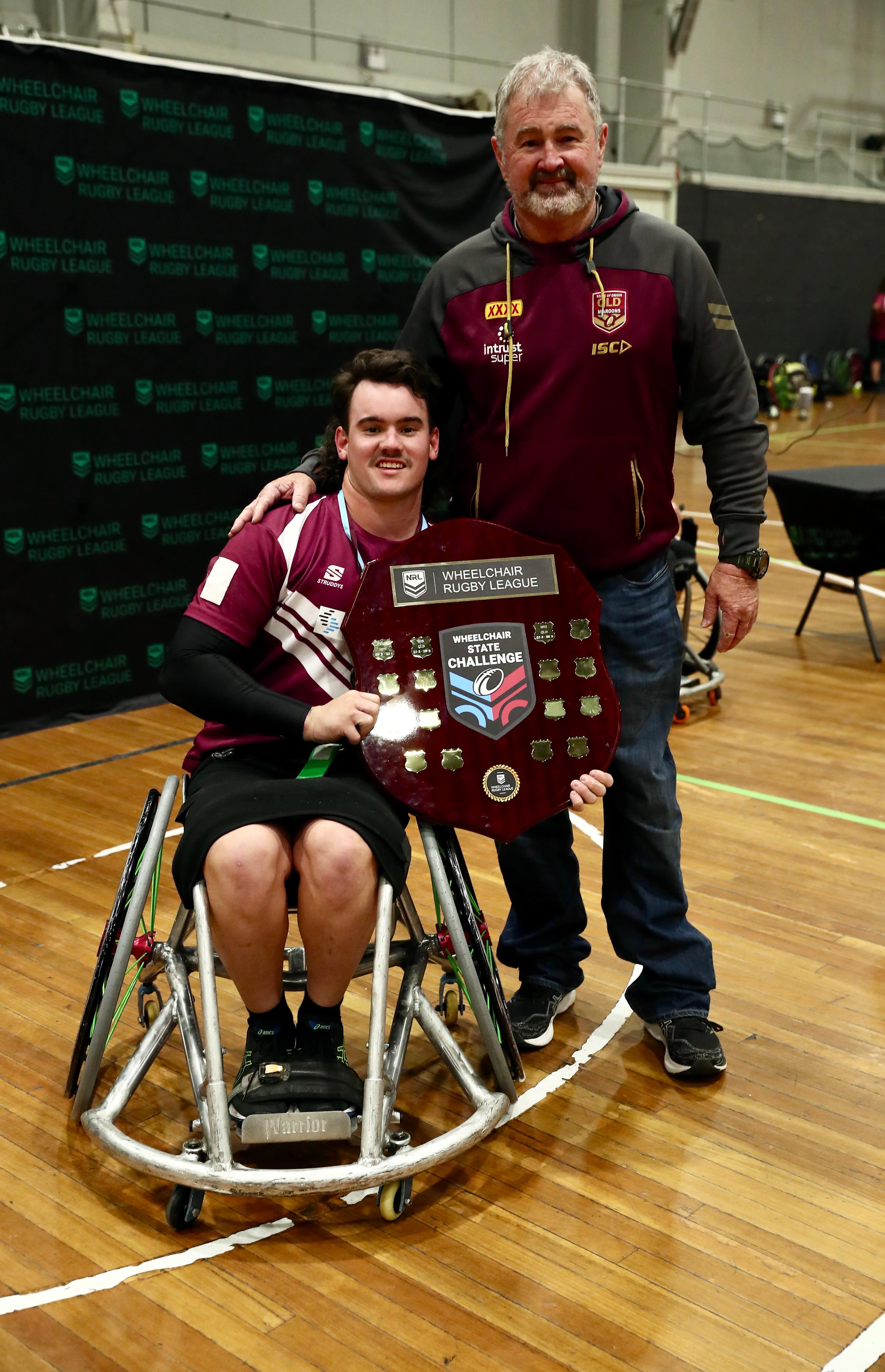 One man sits in a wheelchair holding a shield while another man stands at his back with hand on him