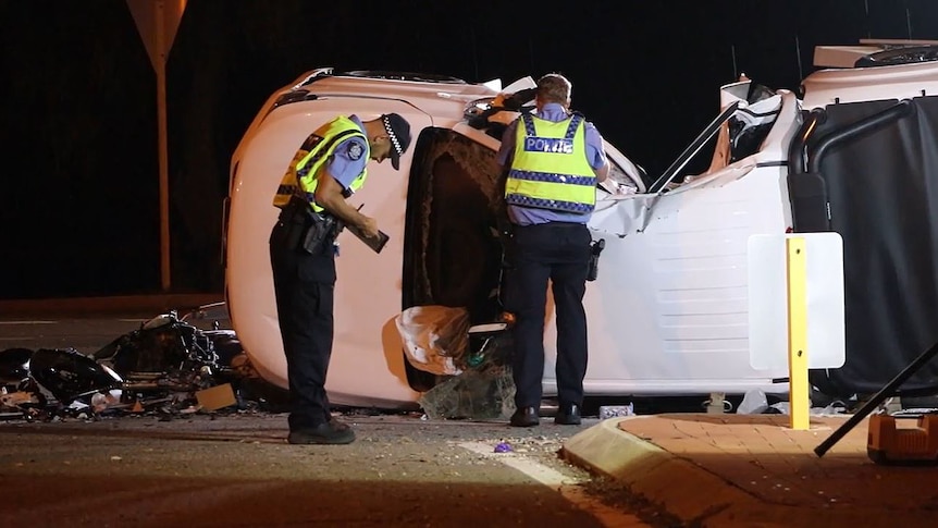 Two police officers in hi-vis vests stand next to a white ute lying on its side at a crash scene, near a wrecked motorbike.
