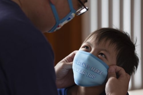 An adult helps a young boy fit a face mask that reads: My parents make me wear this.