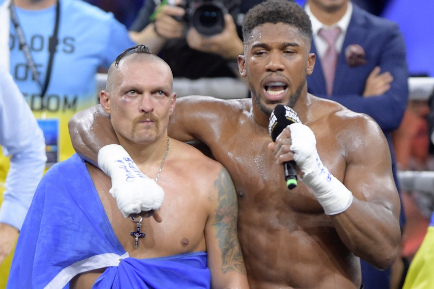 Anthony Joshua talks down a microphone with his arm around an unimpressed Oleksandr Usyk