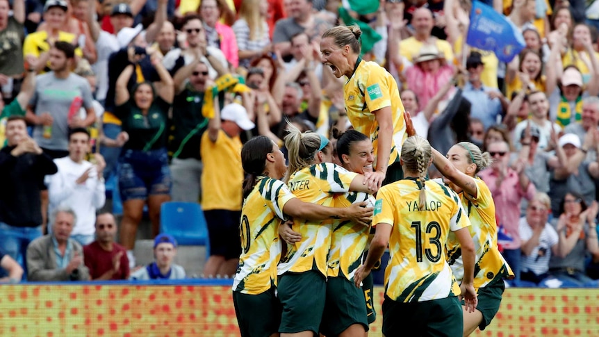 The Matildas huddle in celebration in front of a roaring crowd