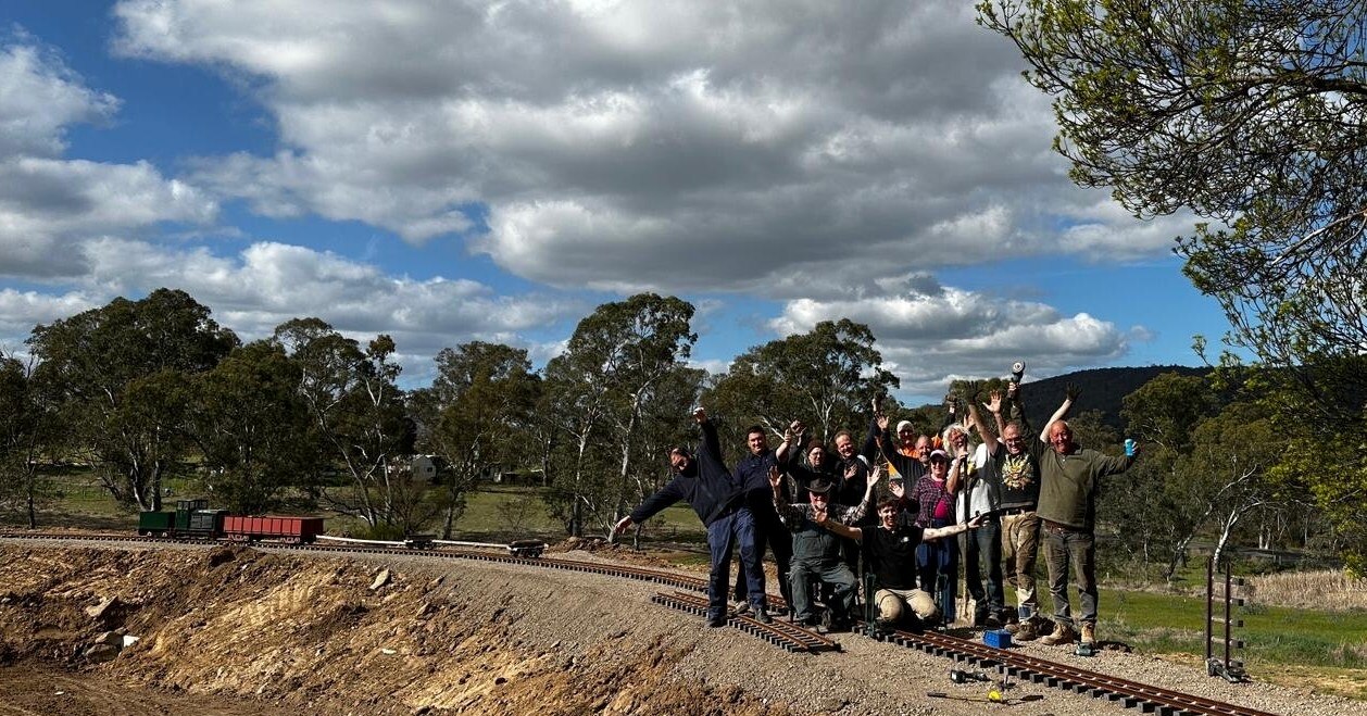 Un grupo de personas se encuentra en una vía de tren en miniatura frente a un matorral.
