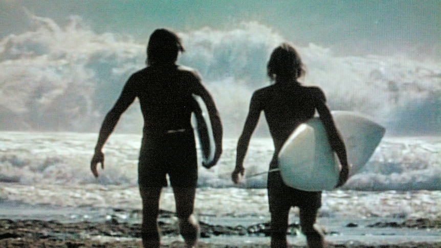 Two silhouettes stand facing of rough surf. The figure on the right holds a surfboard. 