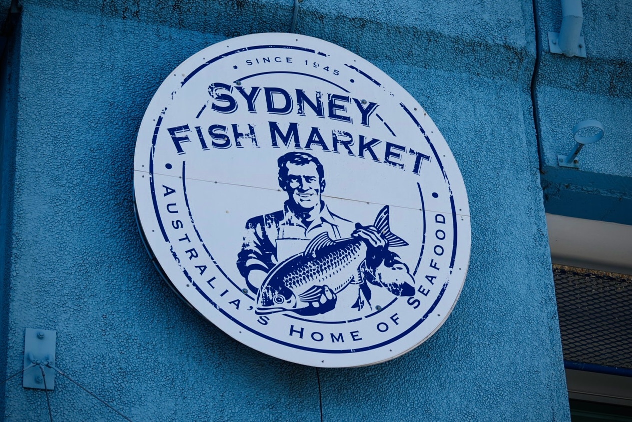 Um logotipo azul e branco do Mercado de Peixe de Sydney mostrando um homem segurando um peixe.