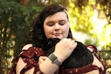 A woman hugs her dark-furred rabbit while sitting outside.