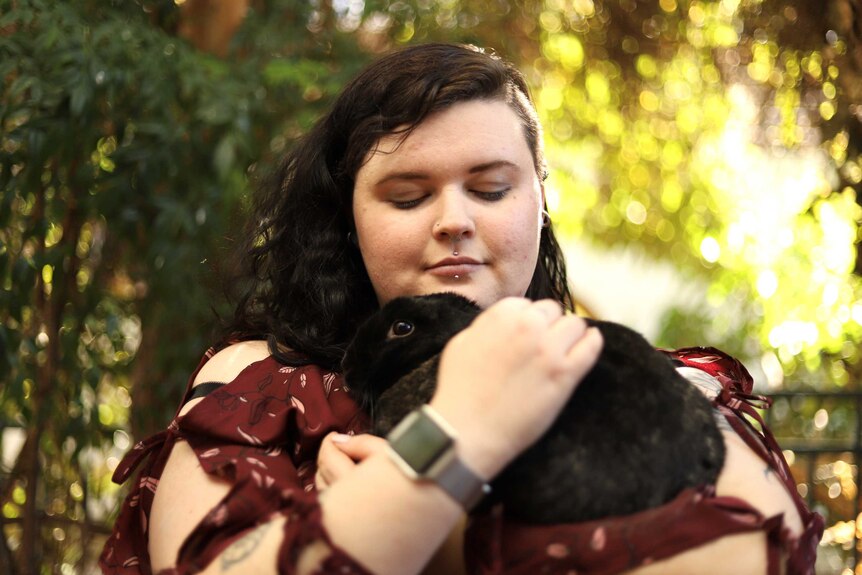A woman hugs her dark-furred rabbit while sitting outside.