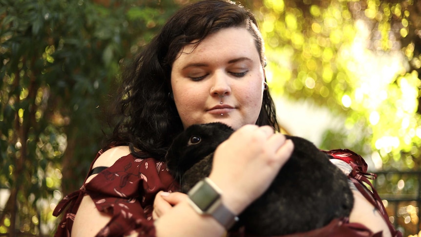 A woman hugs her dark-furred rabbit while sitting outside.