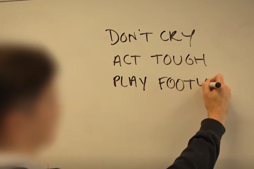 A teenage boy whose face is blurred writes phrases on a whiteboard: Don't cry, act tough, play footy.