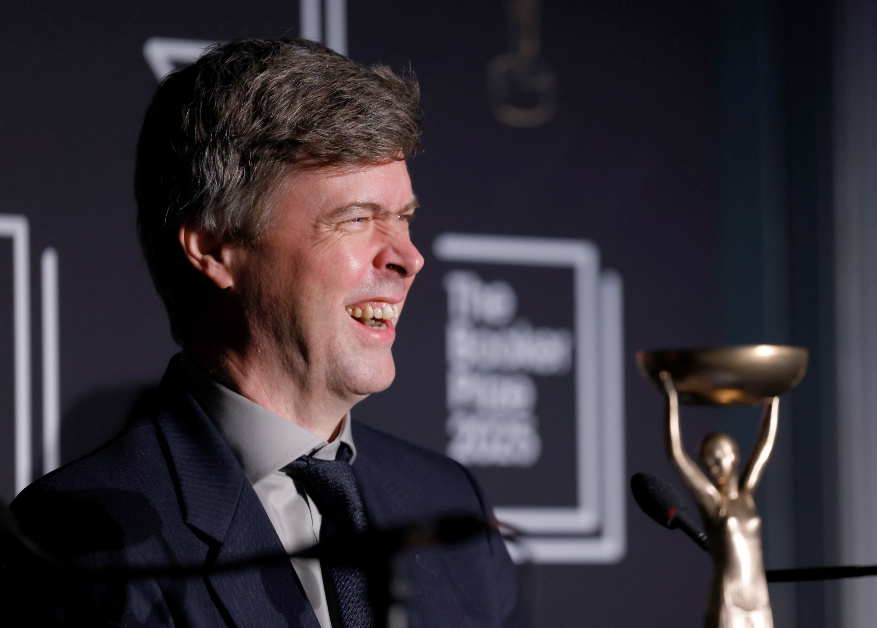 A man with short dark grey hair, in a suit and tie, smiling with his nose scrunched up, with a trophy in the background