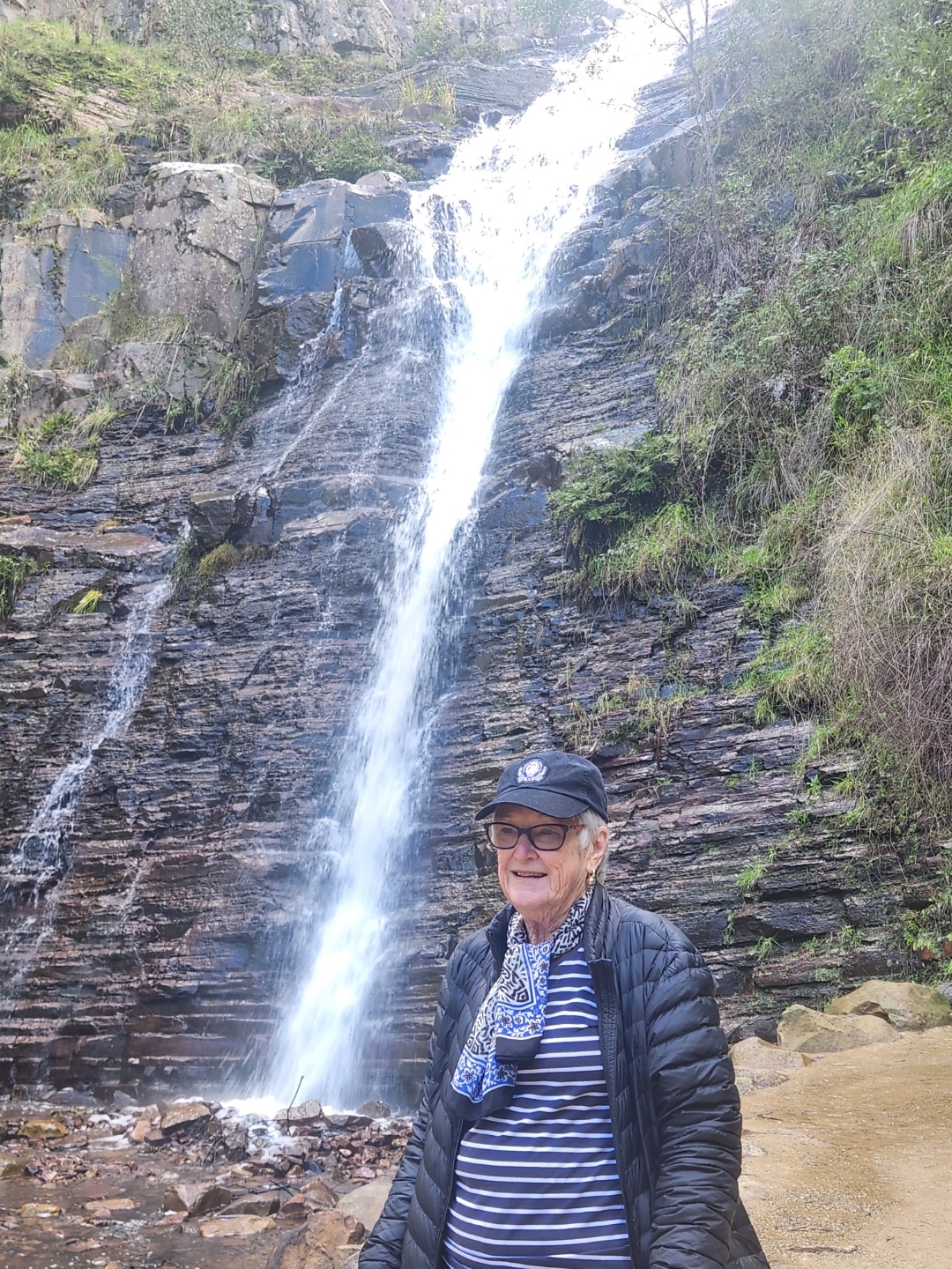 A woman in a cap posed in from of a tall, narrow waterfall.