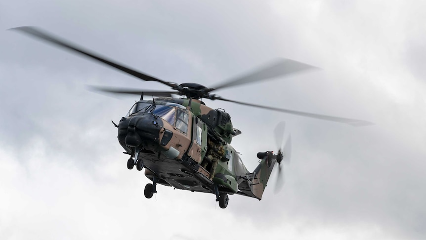 A green and brown camouflage coloured helicopter flies through the air.