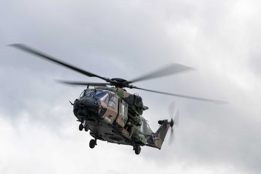 A green and brown camouflage coloured helicopter flies through the air.