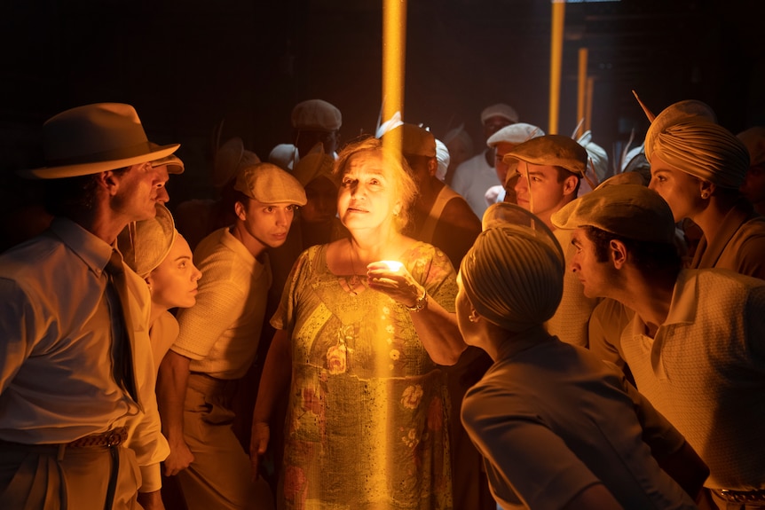 An older latina woman holds a candle, surrounded by a group of people at night, in the film In The Heights 