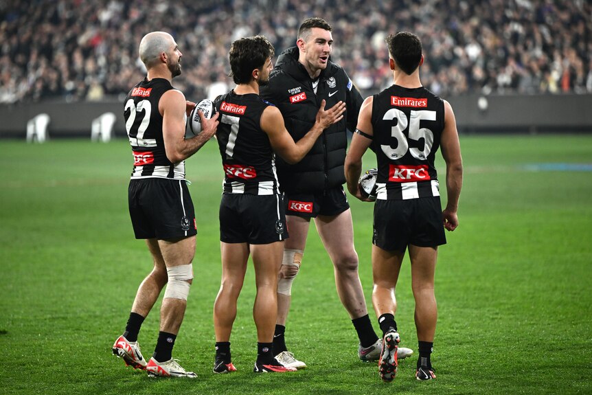 Three Collingwood players gather around an injured teammate who is wearing his team jacket after a finals win.