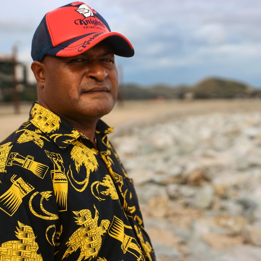 A man in a cap and tropical shirt stands on a beach 