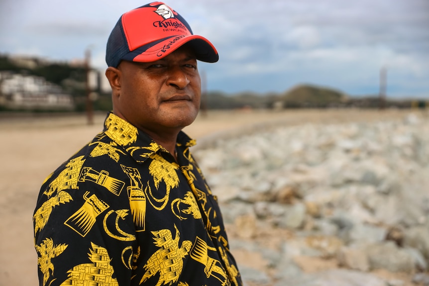 A man in a cap and tropical shirt stands on a beach 
