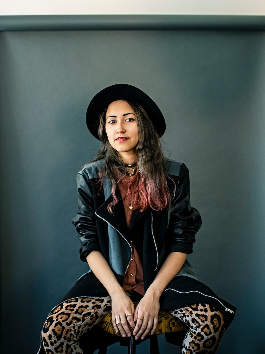 Anchuli Felicia King wears black hat and jacket, leopard patterned pants, sits on stool in front of grey background.