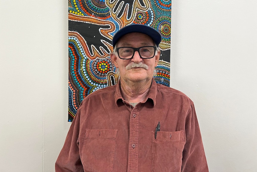 An elderly man stands smiling at camera in front of a painting. 