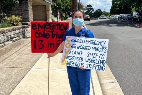 A woman in nursing scrubs and a mask holds protest signs