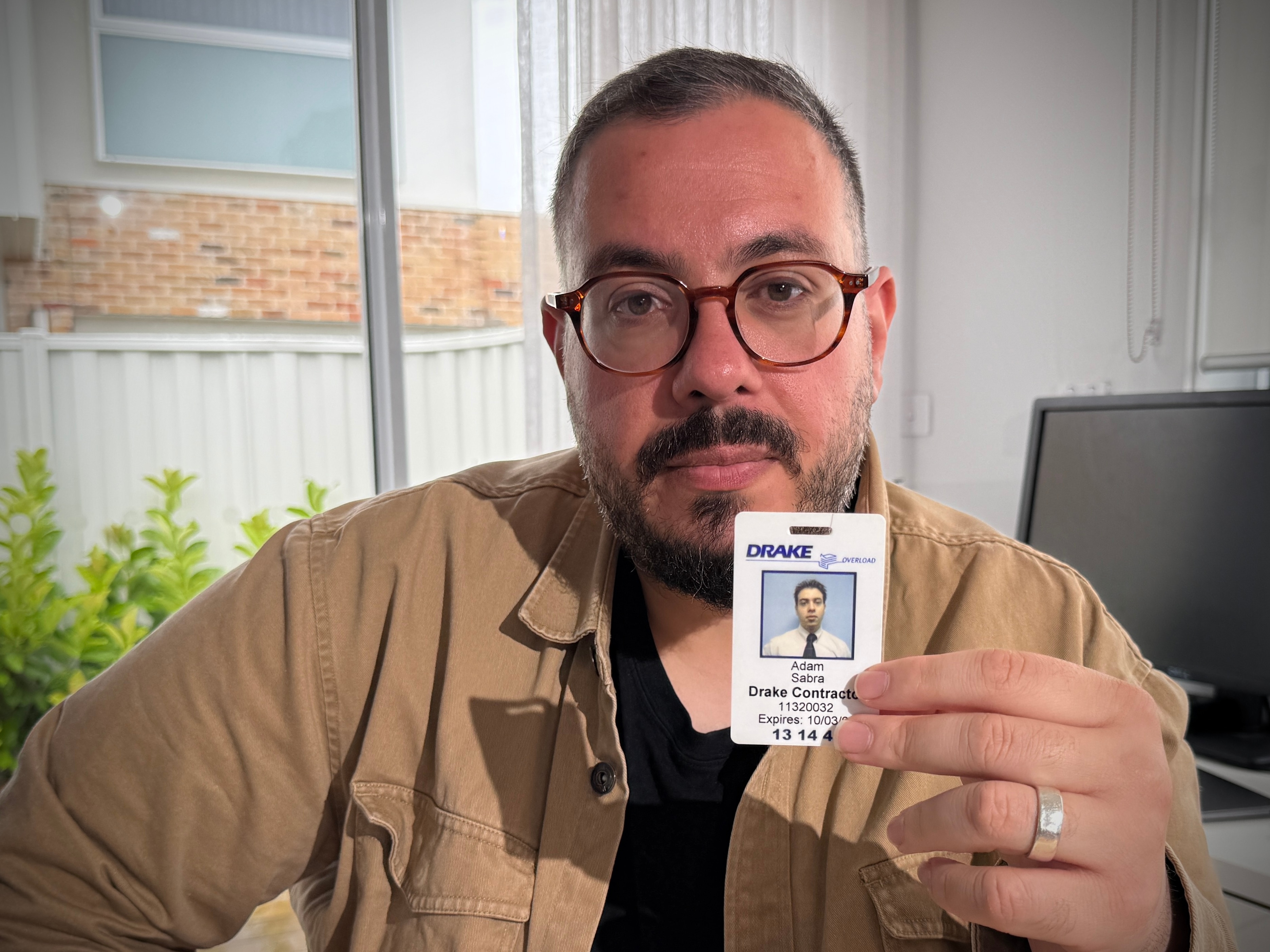 A man wearing glasses sits at a desk and holds up his old ID which has the anglicised name 'Adam' on it.