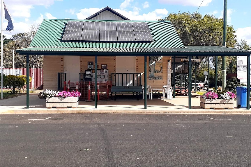 A weatherboard building with a green roof and solar panels provides shade for a bench, community notice board and damaged ATM.