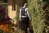 A police officer in a protective vest stands at the front of a house with trees and plants either side.