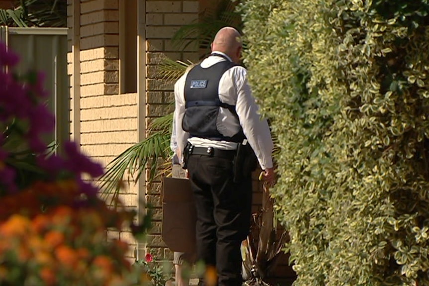 A police officer in a protective vest stands at the front of a house with trees and plants either side.